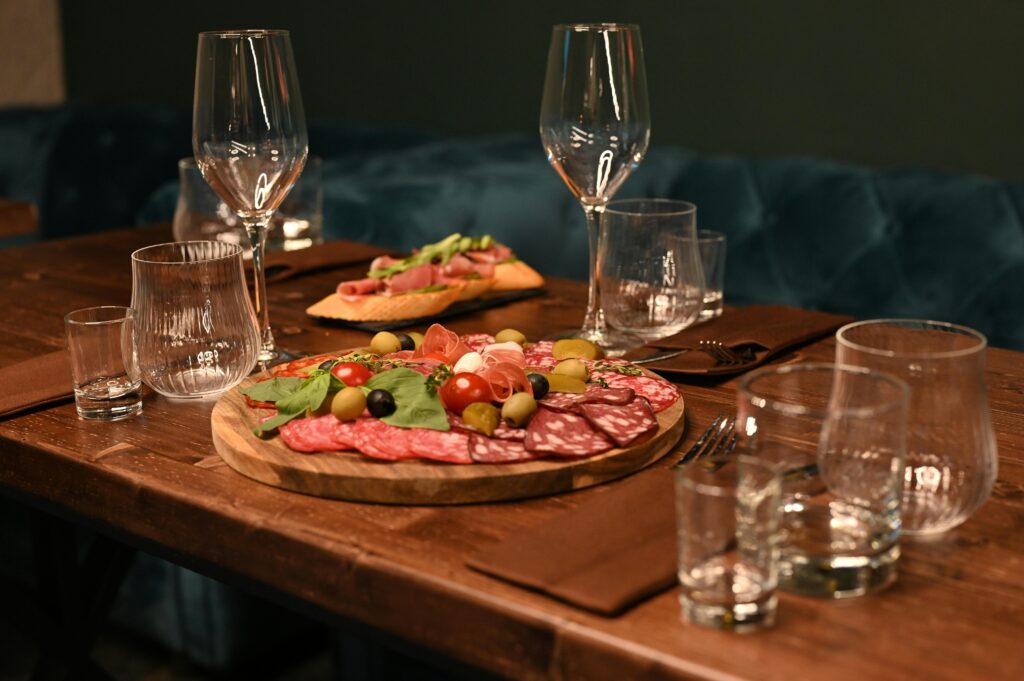 Rustic table with various cured meats such as chouriço, linguiça, and salami, accompanied by artisanal bread and a bottle of wine in the background